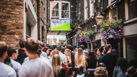A large crowd watching live football on an outdoor screen in an alley near St Christopher's Pub, Borough High Street